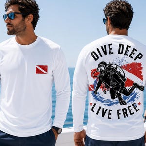 May include: A white long-sleeve shirt featuring a small red and white diver down flag on the front chest. The back displays a graphic of a scuba diver with the text DIVE DEEP LIVE FREE, surrounded by a red and blue circular brushstroke design.