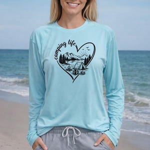 Puede incluir: Camiseta de manga larga azul claro con un gráfico negro en forma de corazón que dice "camping life". El gráfico presenta una escena de montaña con una tienda de campaña, árboles y un sol. La persona lleva pantalones cortos grises.