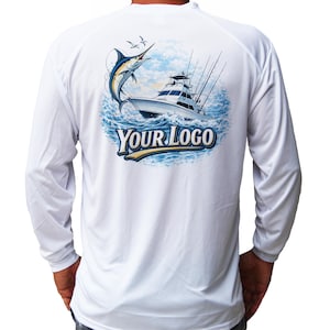 Op de afbeelding: Wit shirt met lange mouwen met een afbeelding van een marlijn die uit de zee springt in de buurt van een vissersboot. Het ontwerp bevat de tekst "YOUR LOGO" in blauw en goud. Het shirt is gemaakt van licht materiaal.