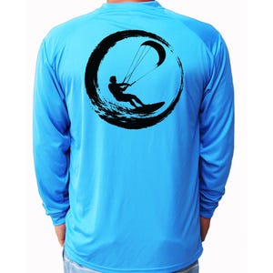 May include: A bright blue long-sleeve shirt featuring a black graphic on the back. The design depicts a silhouette of a person kiteboarding, framed within a circular, brushstroke-style border that resembles a wave or an abstract ring shape.