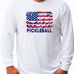 May include: White long-sleeve shirt with a graphic of an American flag made of pickleball paddles and the word "PICKLEBALL" in blue lettering.