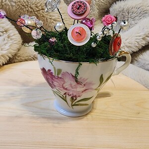 May include: A decorative teacup filled with green moss and adorned with various embellishments. The arrangement includes pink buttons, faux pearls, and crystal-like beads. The teacup features a floral design with pink flowers and gold trim.