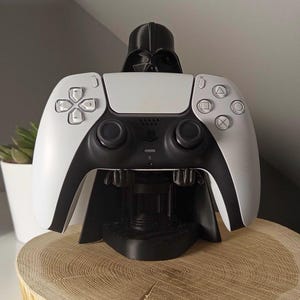 May include: A white and black PlayStation 5 controller rests on a black Darth Vader-themed charging station. The controller is on a wooden slice, with a small plant in a white pot in the background. The controller has a black center and white sides.