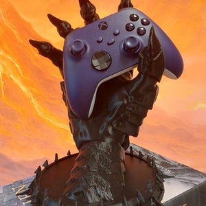 May include: A black, stylized controller stand shaped like a claw holding a purple Xbox controller. The stand has a circular base with sharp edges. The controller has multiple buttons and joysticks. The background is an orange and yellow gradient.
