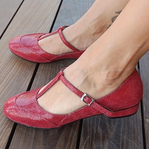 May include: Red leather Mary Jane style shoes with a textured finish. The shoes have a T-strap design with a small buckle closure and a low heel. The shoes are displayed on a wooden surface.
