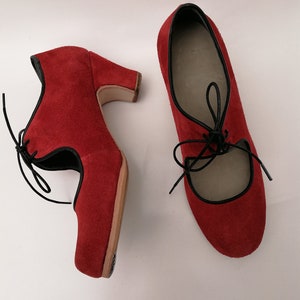 May include: A pair of red suede shoes with black trim and laces. One shoe has a unique heel design, while the other is a classic Mary Jane style. The shoes are set against a white background, showcasing their vibrant color and design.