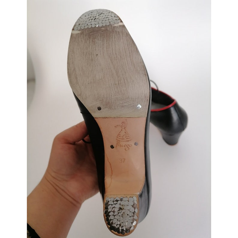 May include: Black flamenco dance shoes with a red trim. The soles are worn, showing the wear and tear from use. The heel is approximately 2 inches high. The brand name "Fuego" is visible on the sole.