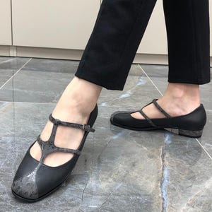 May include: Black T-strap shoes featuring a textured leather finish, a rounded toe, and a low block heel. The design includes a contrasting grey patterned panel on the toe and along the T-strap, secured with a thin adjustable buckle strap.