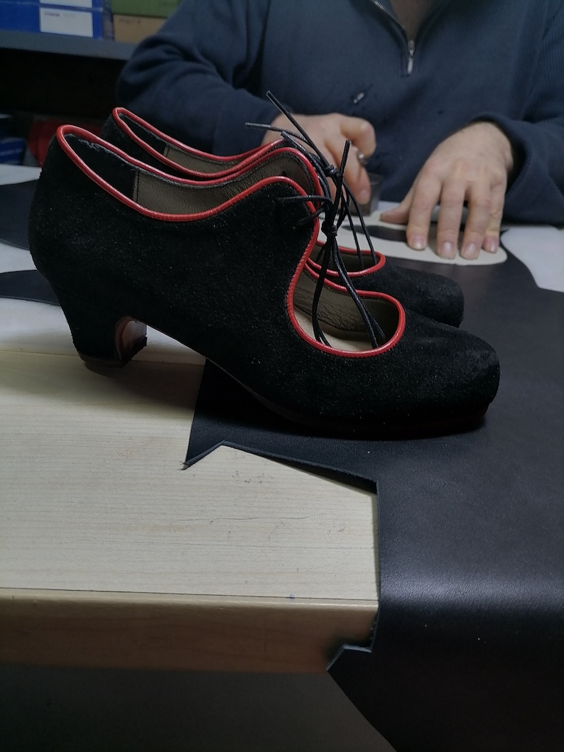 May include: A pair of black suede shoes with red trim and laces. The shoes have a rounded toe and a small heel. The shoes are sitting on a light-colored wooden surface.