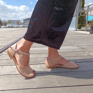 May include: Beige leather sandals with a T-strap design. The sandals feature a textured pattern on the toe box and a buckle closure. The soles are flat and the sandals are open-toed.