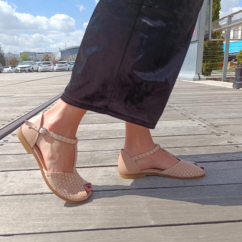 May include: Beige leather sandals with a T-strap design. The sandals feature a textured pattern on the toe box and a buckle closure. The soles are flat and the sandals are open-toed.