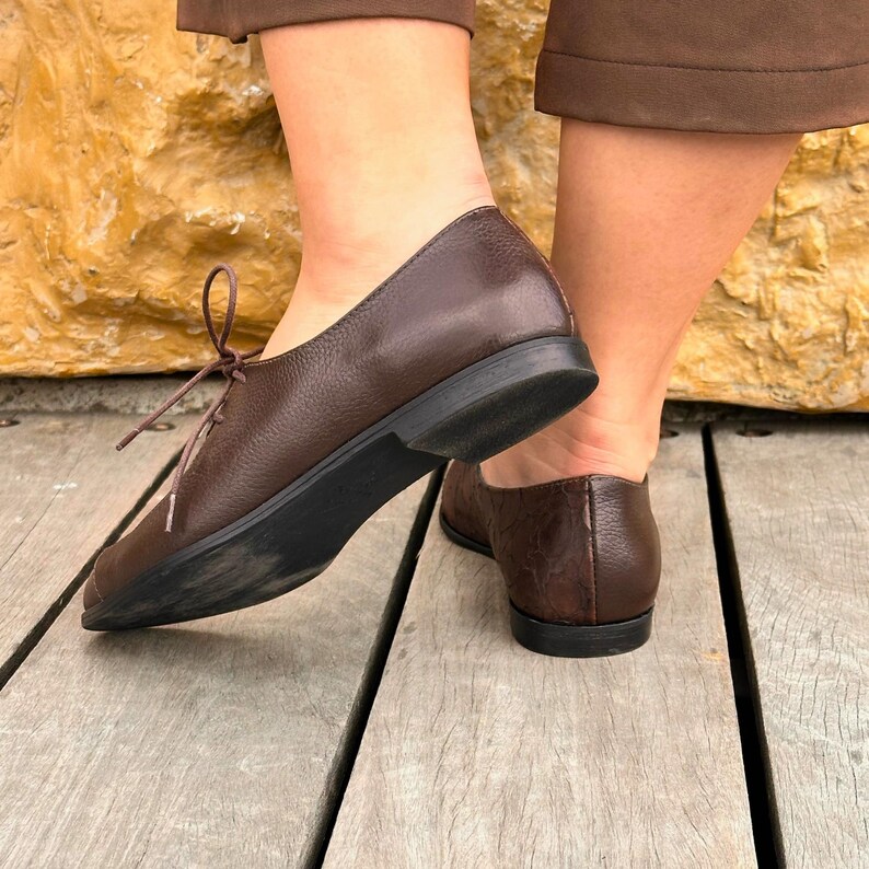 May include: A pair of brown leather lace-up shoes with a rounded toe and a low heel. The shoes are worn by a person standing on a wooden surface.