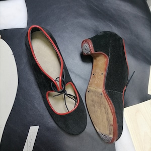 May include: Black suede shoes with red trim and laces. The shoes have a low heel and a cutout design on the top. The soles are a light brown color.
