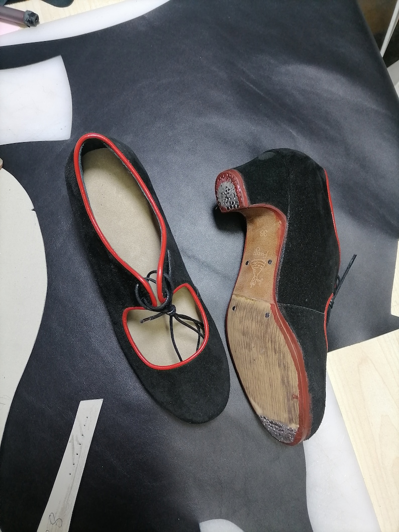 May include: Black suede shoes with red trim and laces. The shoes have a low heel and a cutout design on the top. The soles are a light brown color.