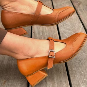 Könnte beinhalten: Ein Paar braune Mary Jane-Schuhe aus Leder mit quadratischer Spitze und einem Riemen mit Schnalle an der Seite. Die Schuhe haben einen kleinen Blockabsatz. Die Schuhe haben eine warme, hellbraune Farbe.