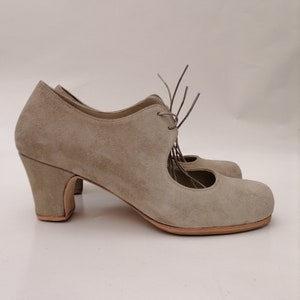 May include: A pair of light taupe suede heeled shoes. The shoes have a rounded toe, a mid-height block heel, and a lace-up closure. The design includes a cutout detail on the side. The soles are a light brown color.