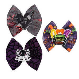 May include: Three Halloween-themed hair bows. One gray bow features a heart with a knife and the words "HORROR MOVIES." Another bow is purple with a spider and web design. The third bow has a purple background with the words "I SMELL CHILDREN."