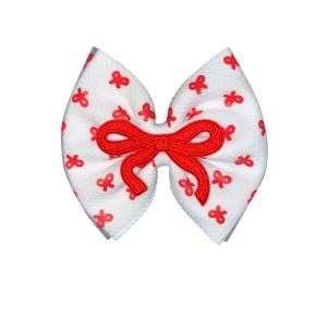 May include: White fabric bow with a red bow design in the centre. The bow is decorated with small red bow shapes scattered across the surface. The bow is a hair accessory.