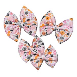May include: Four fabric hair bows with a Halloween theme. The bows are white with a pattern of pumpkins, spiderwebs, flowers, and geometric shapes in orange, pink, and black. The bows are tied in the center with a matching fabric.