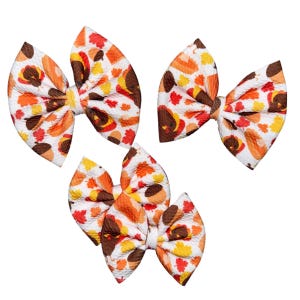 May include: Three white fabric hair bows with a Thanksgiving-themed pattern. The bows feature a repeating design of turkeys, acorns, and fall leaves in shades of brown, orange, and yellow. The bows are tied with a white knot.
