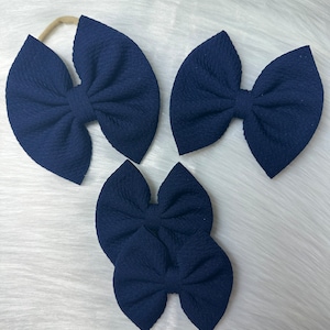 May include: Three navy blue hair bows, one with a headband, two with alligator clips. The bows are made of a textured fabric and are tied in a classic bow shape.