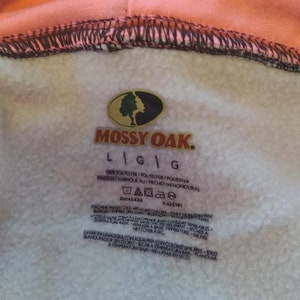 May include: A white care tag with black text for a Mossy Oak brand sweatshirt. The tag includes washing instructions in both English and Spanish. The tag says "100% Polyester/Polyester/Polyester Made in/Fabrique au/Hecho en Honduras RN145424 CA25181"
