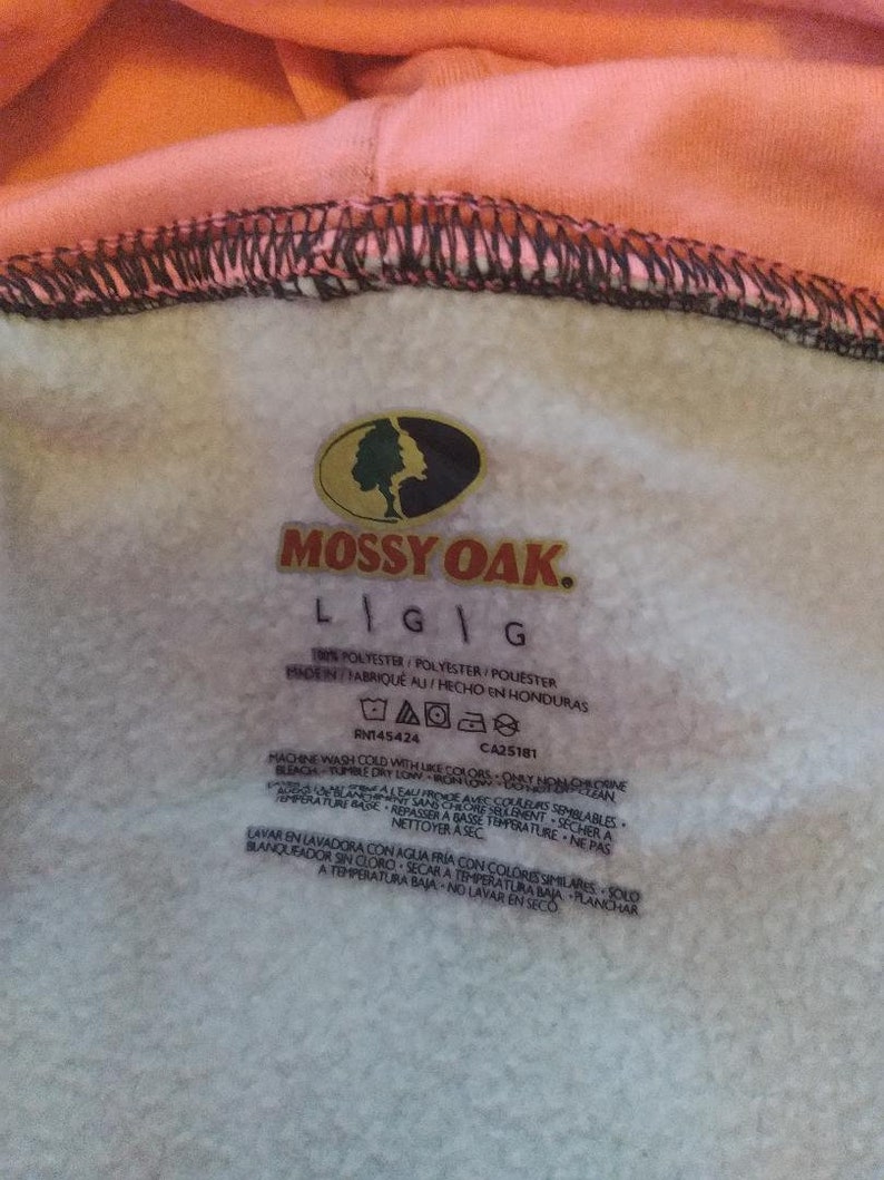 May include: A white care tag with black text for a Mossy Oak brand sweatshirt. The tag includes washing instructions in both English and Spanish. The tag says "100% Polyester/Polyester/Polyester Made in/Fabrique au/Hecho en Honduras RN145424 CA25181"