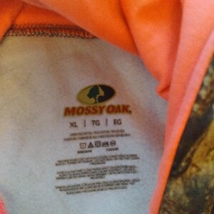 May include: A white tag with the Mossy Oak logo and text. The tag has care instructions and size information, including XL, TG, and EG. The tag is attached to a pink and camouflage patterned hooded sweatshirt.