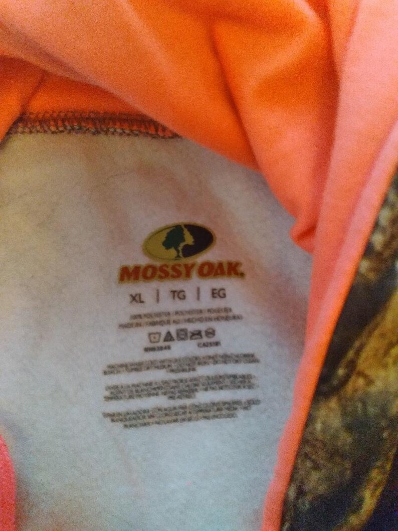 May include: A white tag with the Mossy Oak logo and text. The tag has care instructions and size information, including XL, TG, and EG. The tag is attached to a pink and camouflage patterned hooded sweatshirt.