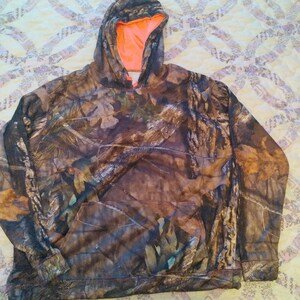 May include: Camouflage hoodie with an orange lining. The hoodie has a drawstring hood and a kangaroo pocket.