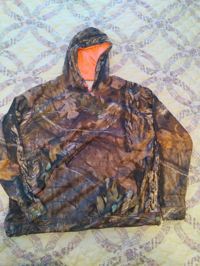 May include: Camouflage hoodie with an orange lining. The hoodie has a drawstring hood and a kangaroo pocket.