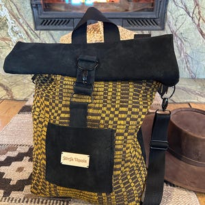 Könnte beinhalten: Ein schwarz-gelb karierter Rucksack mit schwarzem Überschlag und Griff. Die Tasche hat eine schwarze Tasche mit der Aufschrift "Strefa Tkania". Ein brauner Hut ist im Hintergrund zu sehen.