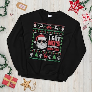 May include: Black Christmas jumper featuring a Santa Claus wearing sunglasses and the text "I got ho's in different area codes".