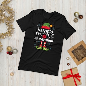 May include: Black t-shirt with a Christmas elf design. The elf is wearing a red hat, green pants, and green shoes. The text on the shirt reads "Santa's Favorite Paramedic".