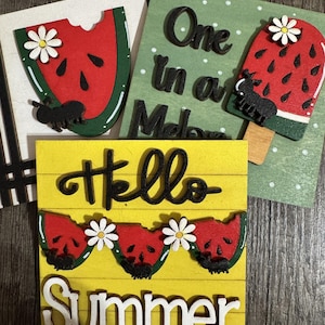 May include: Wooden summer-themed decorations featuring watermelon slices, daisies, and the words "Hello Summer" in black and white. One piece says "One in a Melon". The decorations are on a wooden surface.