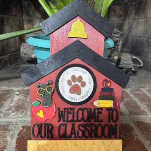 May include: A wooden classroom decoration in the shape of a schoolhouse. It features a red body with a black roof, a yellow bell, and the words "WELCOME TO OUR CLASSROOM." Includes a ruler for scale.