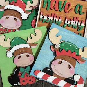 May include: A collection of four Christmas-themed wooden signs featuring cartoon moose characters. The signs are painted in various colors, including green, blue, and brown, and decorated with festive hats, bows, and candy canes. One sign reads "Have a holly jolly."
