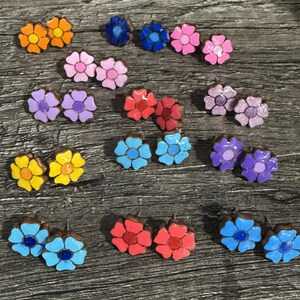 May include: A collection of colorful flower-shaped stud earrings. The earrings feature various colors, including orange, yellow, blue, pink, red, and purple. Each earring has a dark brown base. The earrings are arranged on a wooden surface.