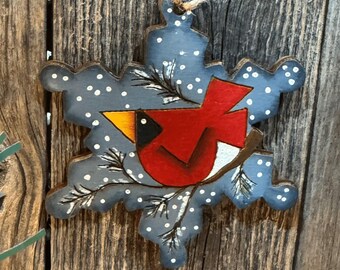May include: A wooden snowflake ornament with a painted cardinal bird design. The snowflake is blue with white polka dots and a natural twine hanger. The cardinal is red with a yellow beak and is perched on a branch with green leaves.
