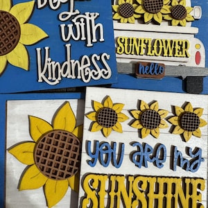 May include: Wooden sunflower-themed signs with inspirational quotes. One sign reads "Begin with Kindness" on a blue background. Another features a white truck with sunflowers and the word "Sunflower". A third sign says "You are my Sunshine".