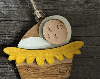 May include: Wooden Christmas ornament depicting a baby in a manger. The baby has closed eyes and a smiling expression. The manger is yellow and brown, with a textured wood grain appearance. A twine loop is attached for hanging.