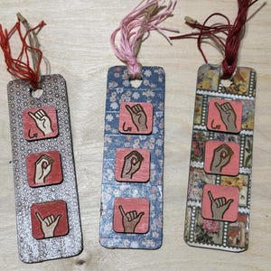 May include: Three handmade bookmarks with different patterned backgrounds. Each bookmark features three small, square wooden tiles with hand sign illustrations. The bookmarks have red, pink, and brown twine attached at the top.