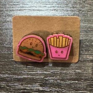 May include: Wooden food-themed earrings featuring a pink-outlined burger and a pink-outlined fries container, both with smiling faces. The earrings are on a brown cardboard backing.