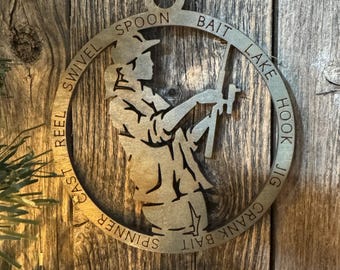 May include: A circular metal ornament featuring a silhouette of a person fishing, holding a fishing rod. The ornament has a ring with the words "REEL", "CAST", "SPINNER", "CRANK BAIT", "JIG", "HOOK", "LAKE", "BAIT", "SPOON", and "SWIVEL".