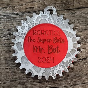 May include: Red and silver ornament with a gear design. It says "Robotics The Super Bots Mr. Bot 2024".