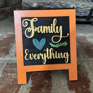 May include: A small, orange-framed sign with the words "Family is Everything" in gold lettering. The sign has a black background with a teal heart and green leaf accents. The sign is freestanding.