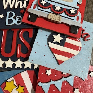 May include: A collection of patriotic wooden decorations in red, white, and blue. The designs include a truck, stars, a heart-shaped American flag, and the letters "US" and "White Be". The truck has the word "USA" on the front.