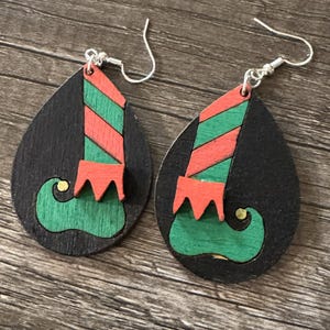 May include: A pair of teardrop-shaped earrings featuring a black background with a painted elf leg design. The elf legs are green and red with a gold accent on the toe. The earrings have silver-colored fish hook ear wires.