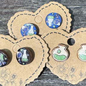 May include: A collection of handmade stud earrings featuring science-themed designs. The earrings are displayed on heart-shaped kraft paper cards. Designs include flasks with green liquid, and blue and white science-themed illustrations.