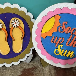 May include: Two decorative wooden coasters. One features yellow flip-flops with orange straps and white flower accents on a purple background. The other says "Soak up the Sun" with a sun graphic.